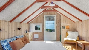 Un dormitorio con techo inclinado, paredes de madera, una cama con ropa de cama blanca, una ventana y una puerta.