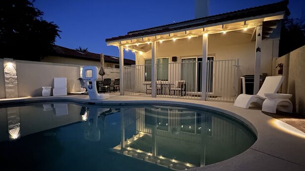 A swimming pool with a covered patio area, lounge chairs, and a small table.