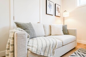 Ein Wohnzimmer mit einer Couch, Kissen, einer Decke, gerahmten Bildern an der Wand und einer Stehlampe.