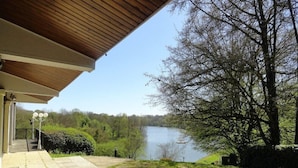 Un espace extérieur abrité offrant une vue sur un lac, un sentier menant à un bâtiment et un paysage bordé d’arbres.