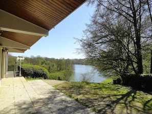 A covered outdoor area with a view of a lake, a pathway leading to a building, and a tree-lined landscape. - Olydea le Domaine du Lambon (Prailles-la-Couarde)