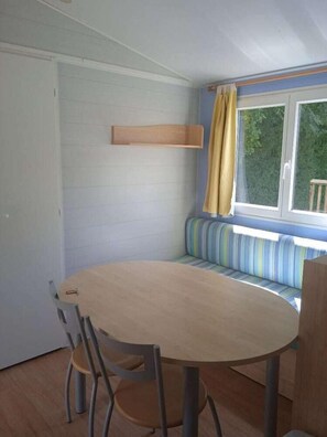 Huoneessa on pyöreä pöytä, tuoleja, tyynyillä varustettu penkki, ikkuna, jossa on verhot, ja puinen hylly.