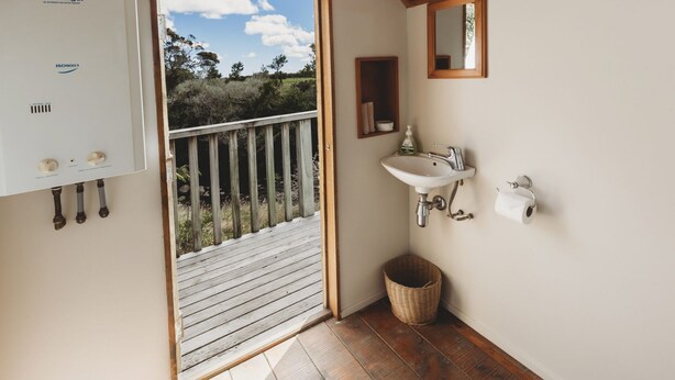 Een badkamer met een wit wastafel, een spiegel en een toiletrolhouder. Er is een houten deur die leidt naar een buitenruimte met een houten terras.
