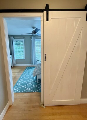 Un couloir avec un plancher en bois, une porte dotée d’une poignée noire et un ventilateur de plafond.