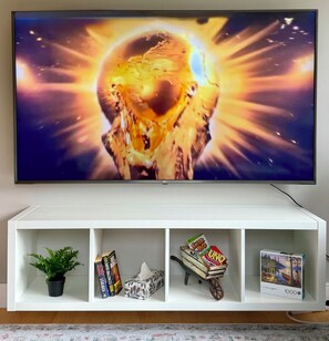 Un televisor de pantalla plana montado en un mueble blanco de entretenimiento con un estante inferior que muestra una planta en maceta, libros, una caja de pañuelos, un carrito de juguetes y un juego de mesa.