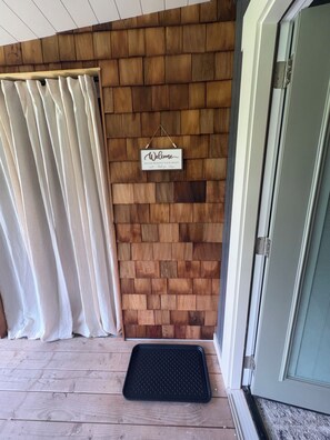 Pared de madera con un letrero de bienvenida, una puerta y un felpudo negro sobre un piso de madera.