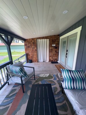 A covered porch with a brick wall, two cushioned chairs, a small table, and a view of a green landscape.