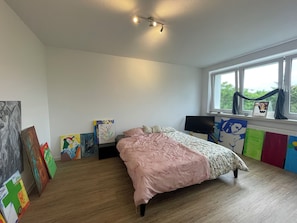 Ein Schlafzimmer mit Bett, Fernseher, Kunstwerken an den Wänden und einem Fenster mit Vorhängen.