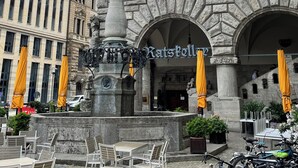 Un edificio storico con una statua, posti a sedere all'aperto e un cartello con su scritto 'KatsKoll'.