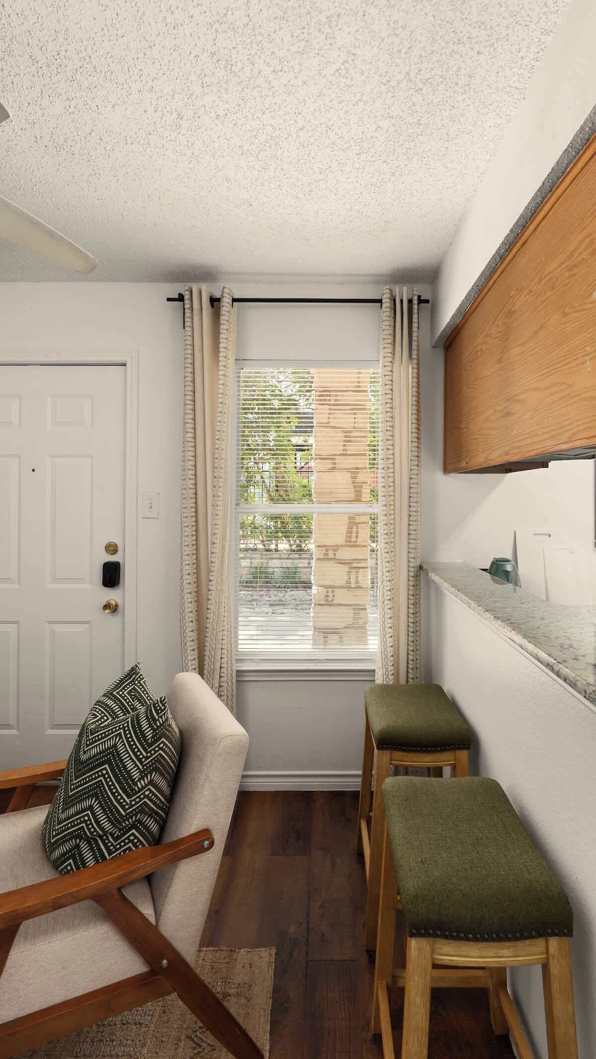 Ein Zimmer mit einer weißen Tür, einem Fenster mit Jalousien, einer hölzernen Bank, einem grünen Hocker und einer Küchenarbeitsplatte.
