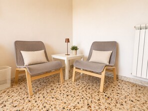 Dos sillones grises con cojines blancos, una mesita pequeña blanca con una lámpara de pie y una planta en maceta, además de un radiador en la pared.