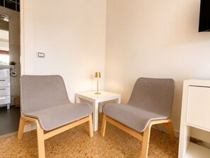 Una stanza con due sedie grigie, un piccolo tavolo bianco con una lampada, un armadietto bianco e un pavimento piastrellato.