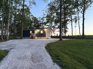 A single-story building with a gravel driveway leading to a wooden door, surrounded by trees and a flag.