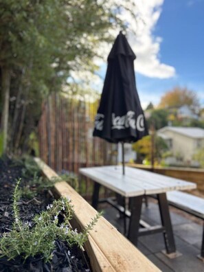 Une terrasse en bois avec une table et un banc, entourée d’un jardin planté et agrémentée d’un parasol noir.