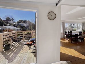 A room with a wooden deck, a dining table with chairs, a clock on the wall, and a view of houses and trees outside.