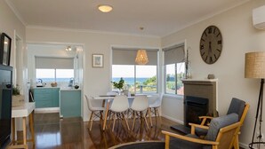 A spacious living area with a fireplace, a dining table with chairs, a kitchen with a view, and a large wall clock.