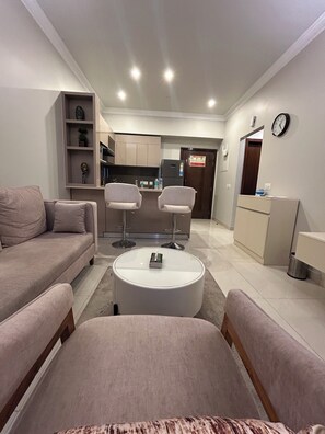 A living room with a sofa, two armchairs, a coffee table, a kitchen area with a refrigerator, and a clock on the wall.