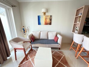 A living room with a sofa, two chairs, a coffee table, a wall-mounted lamp, and a bookshelf.