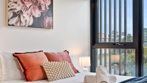 Ein ordentlich bezogenes Bett mit Kissen, ein Fenster mit Jalousien und ein Gemälde an der Wand.
