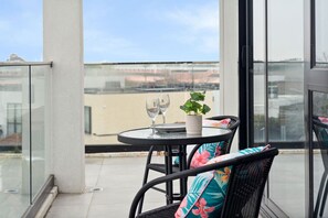 A balcony with a glass railing, a round table with two wine glasses and a potted plant, and wicker chairs with floral cushions. - Frank-2B2B Next To Chadstone (Malvern East)