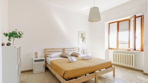Una camera da letto con un letto di legno, pareti bianche, un termosifone, una finestra con tende e un comodino con una lampada, oltre a un quadro appeso al muro.