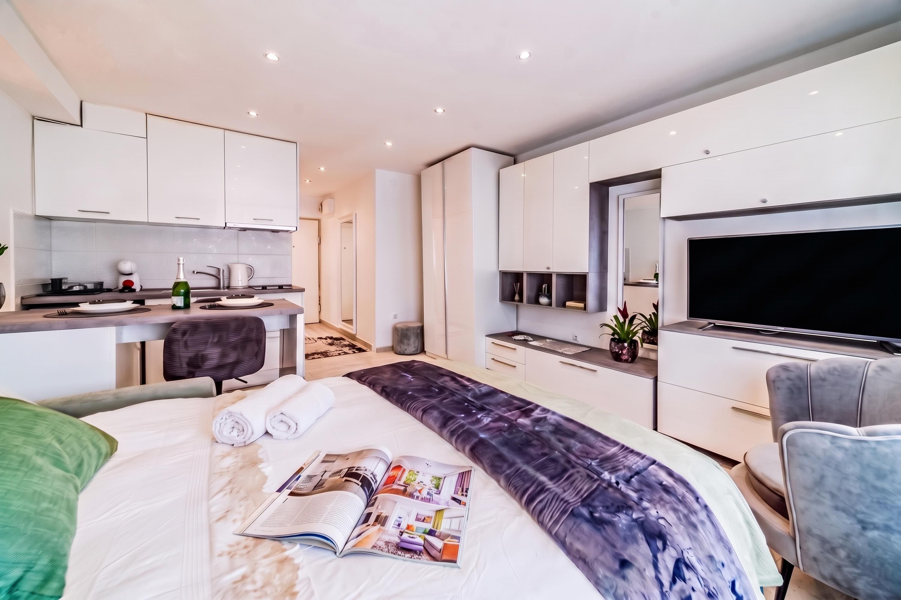 A modern hotel room with a large bed, a flat-screen TV, a dining area, and a kitchen.
