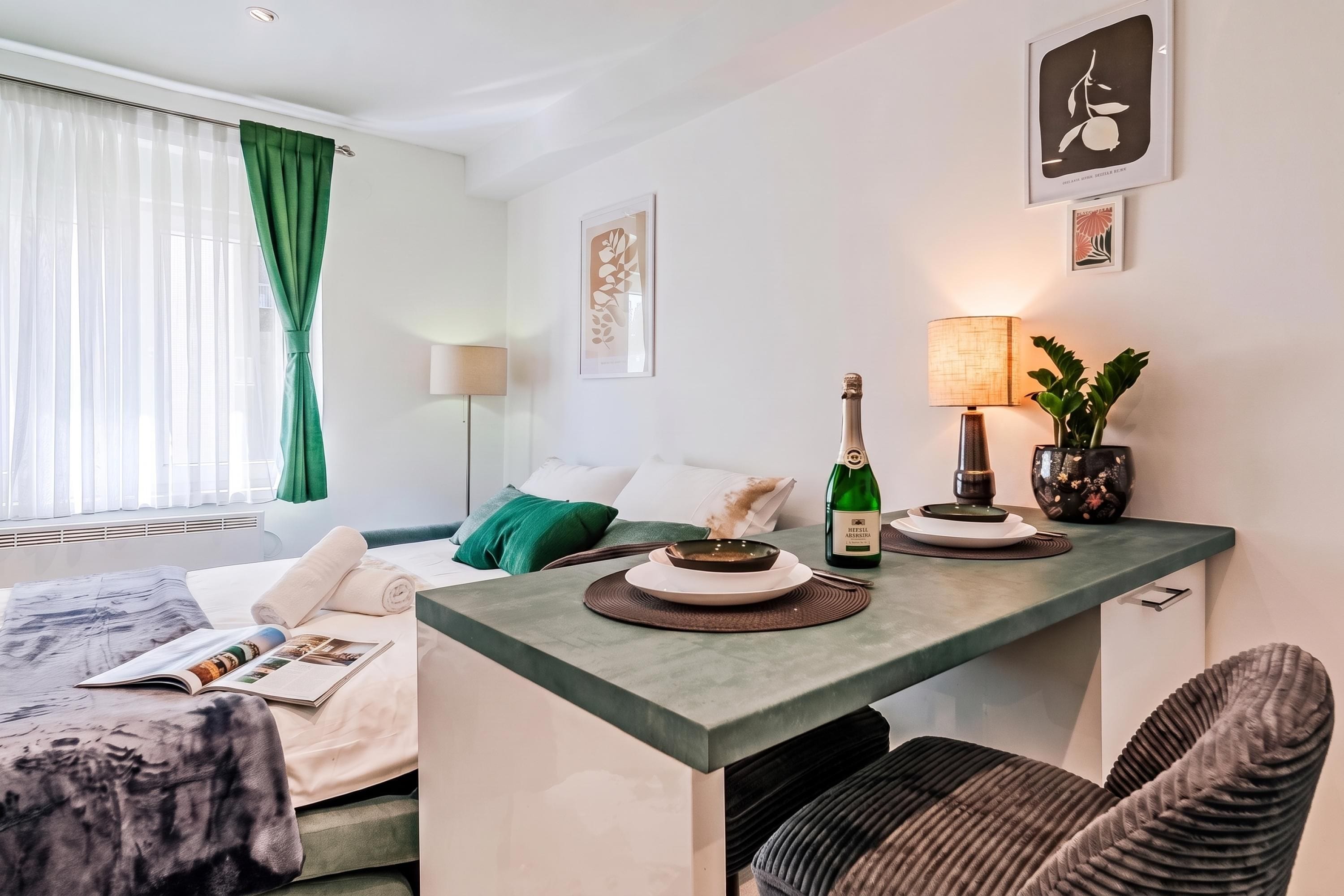 Une chambre d’hôtel avec un lit, une petite table sur laquelle se trouve une bouteille de champagne, une lampe et une plante.