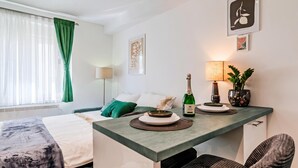Ett hotellrum med en säng, ett litet bord med en flaska champagne, en lampa och en krukväxt.