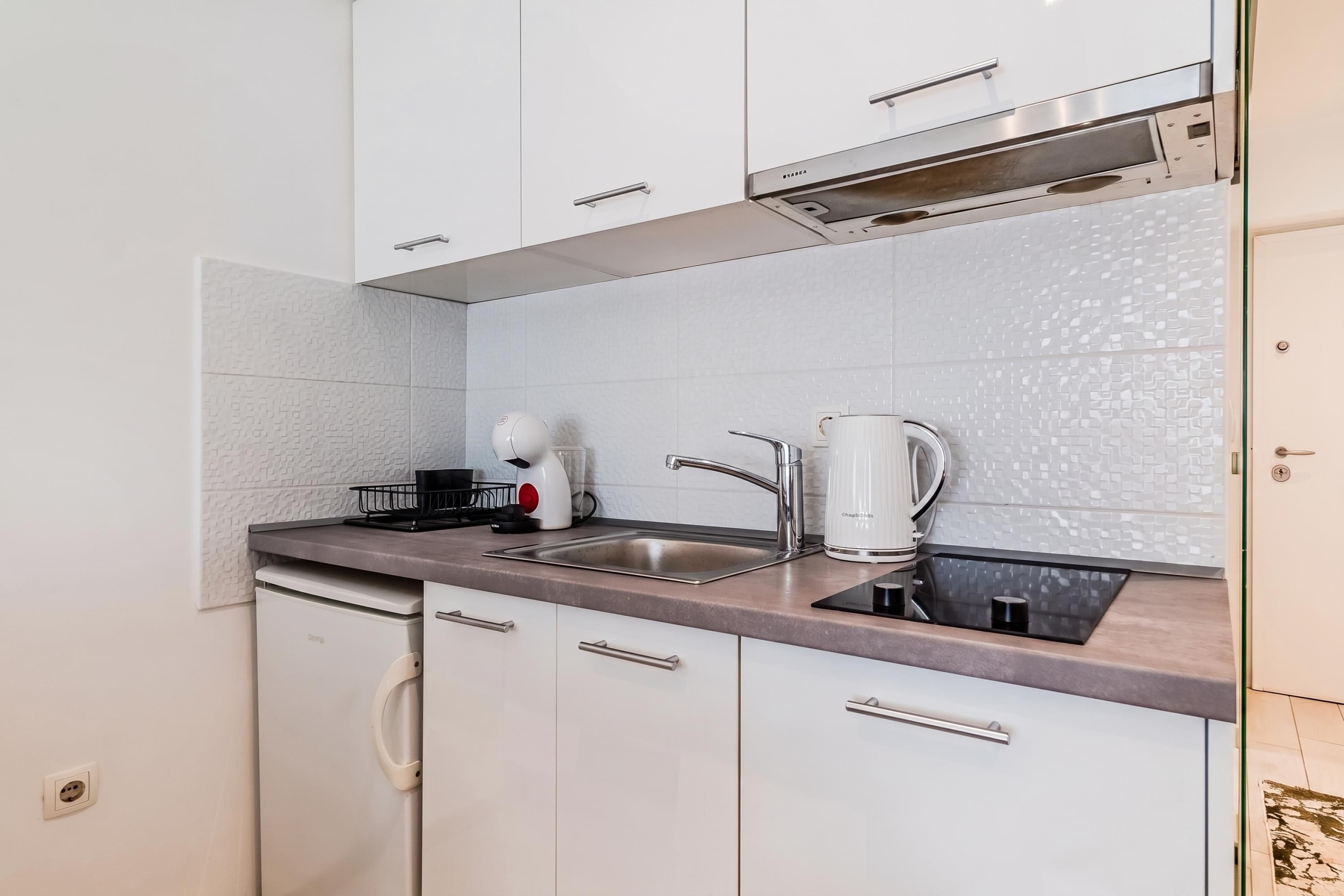 Una cocina compacta con armarios blancos, un lavamanos de acero inoxidable y una cafetera.