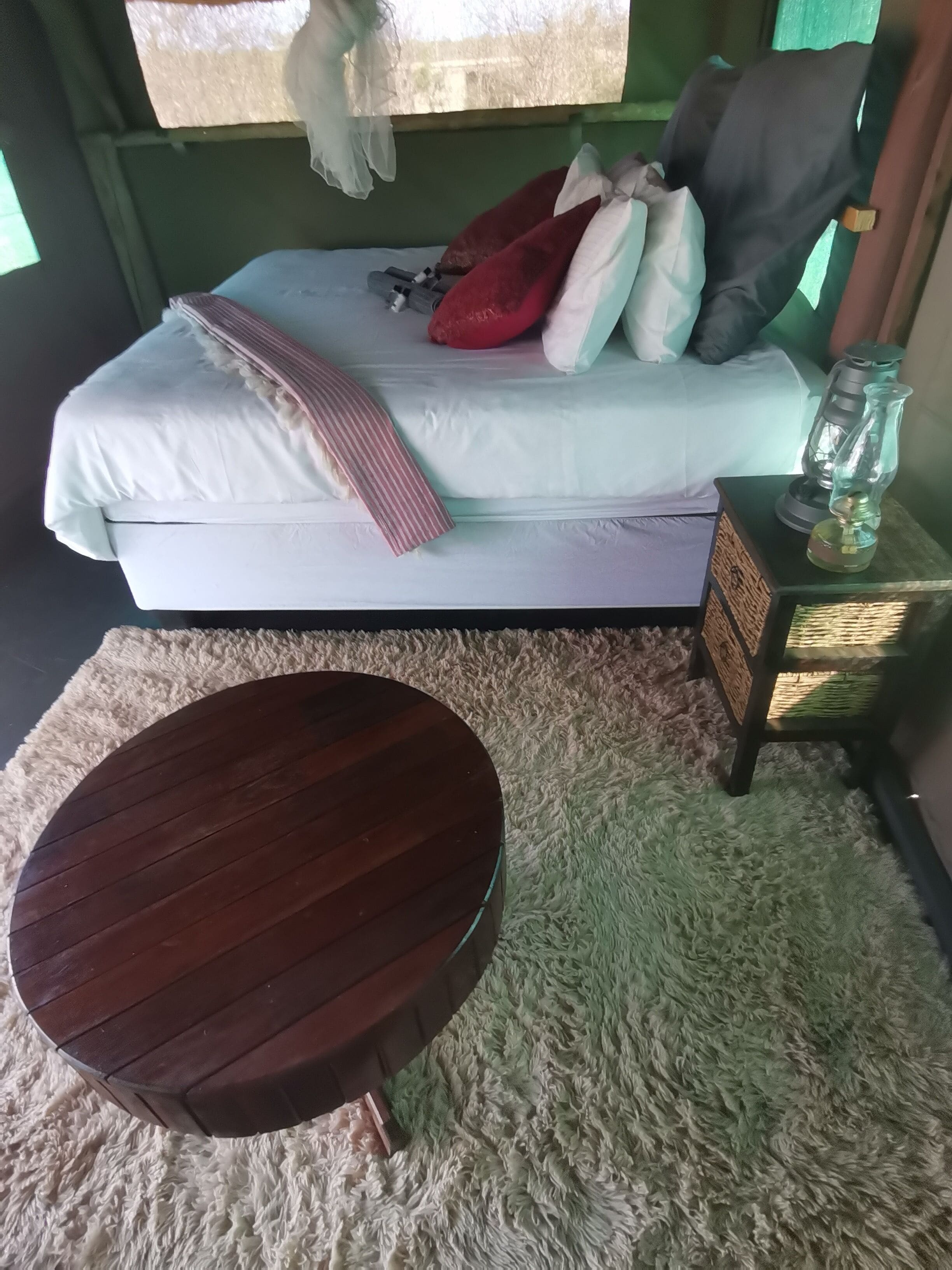 Un letto con cuscini e una coperta, un tavolo rotondo in legno e un comodino con una lanterna.