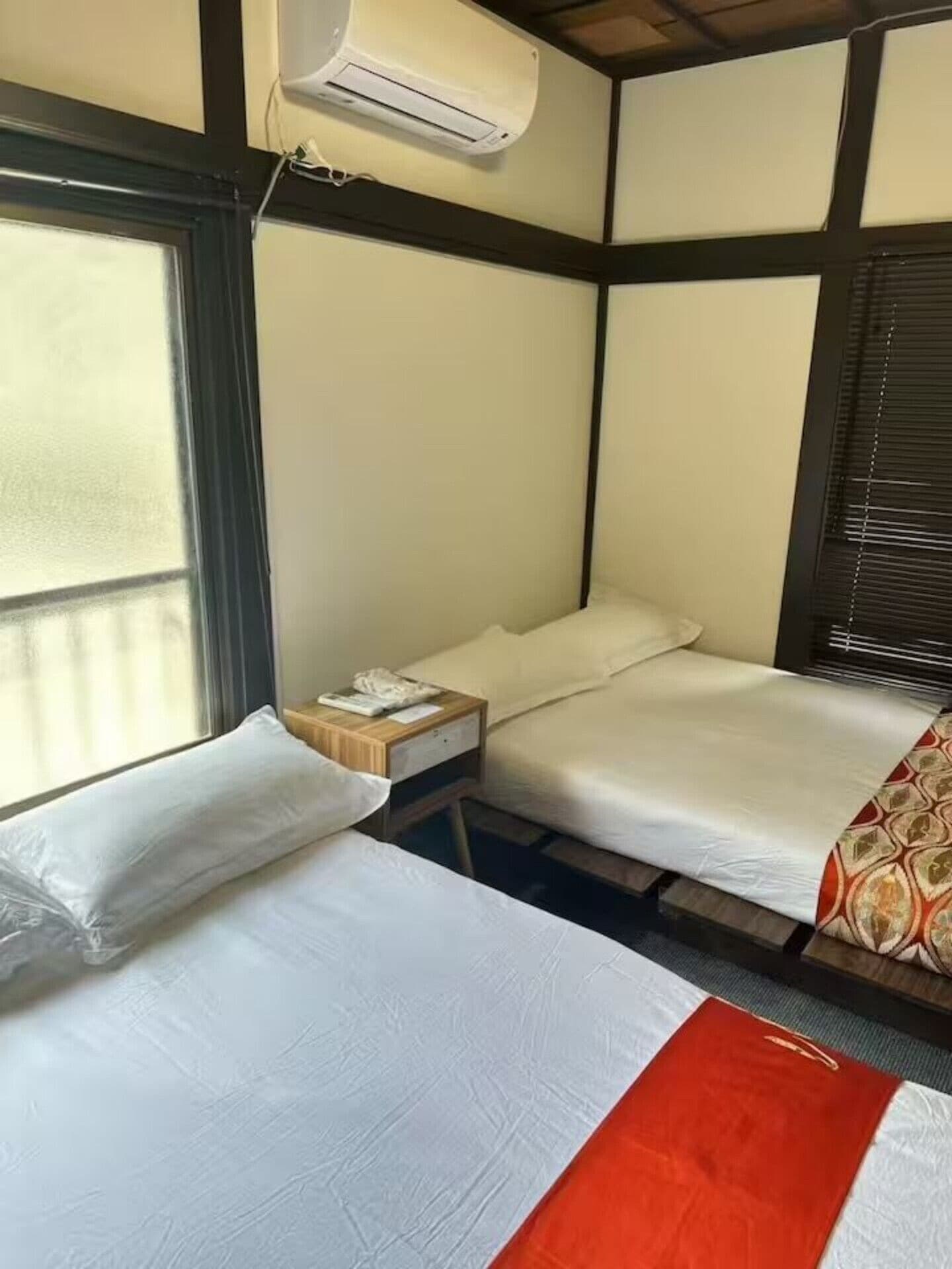 A hotel room with two beds, a window, an air conditioning unit, and a small wooden nightstand.