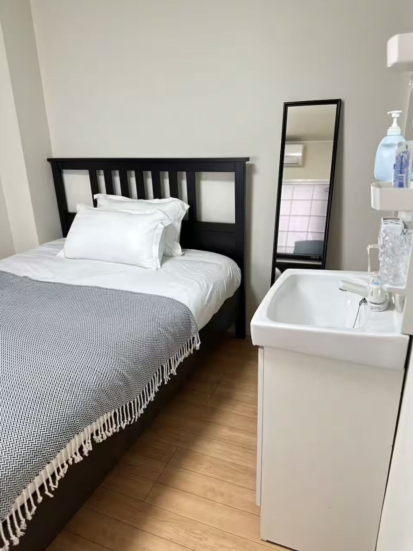 A bedroom with a bed, a bedside mirror, a white sink, and a wooden floor.
