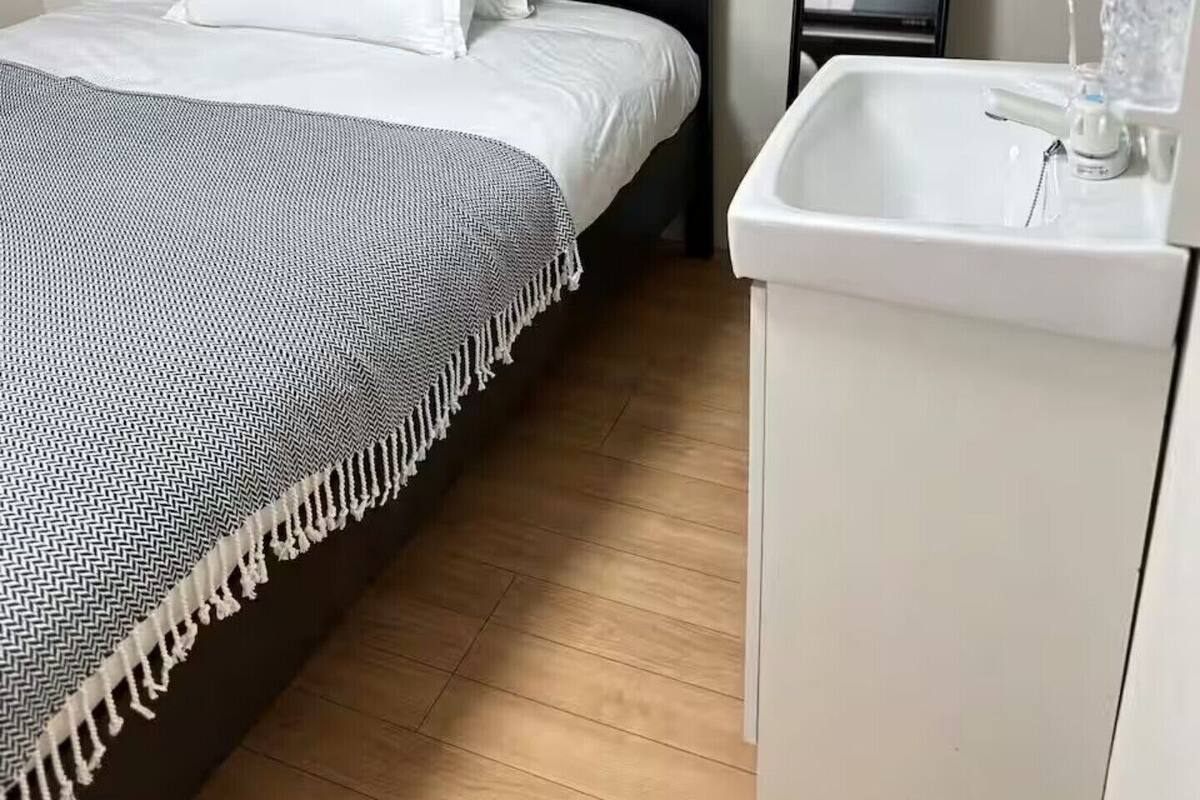 Un dormitorio con una cama, un espejo en la mesita de noche, un lavabo blanco y suelo de madera.