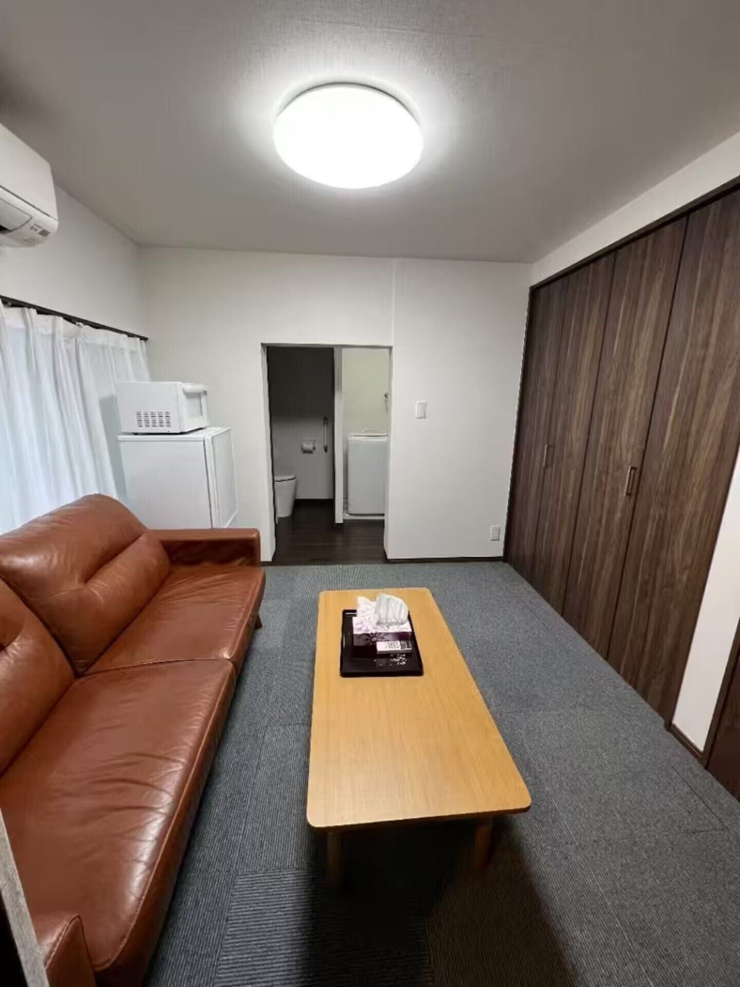 A room with a brown leather sofa, a wooden coffee table, a white refrigerator, a microwave, and a wooden wardrobe.