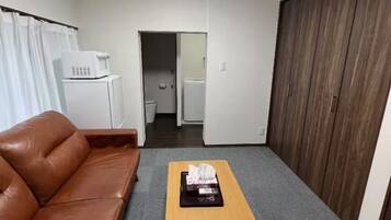 A room with a brown leather sofa, a wooden coffee table, a white refrigerator, a microwave, and a wooden wardrobe.