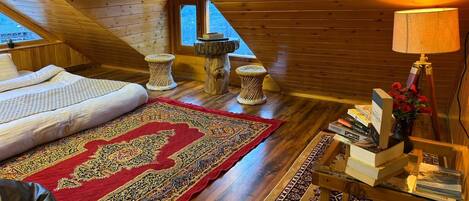 A cozy attic bedroom with a wooden sloped ceiling, a large bed, a red patterned rug, and a small wooden table with books and a lamp.