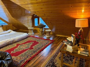 A cozy attic bedroom with a wooden sloped ceiling, a large bed, a red patterned rug, and a small wooden table with books and a lamp.