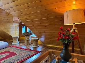 A cozy attic room with wooden slats, a red patterned rug, a vase with red flowers, and a lamp on a tripod.