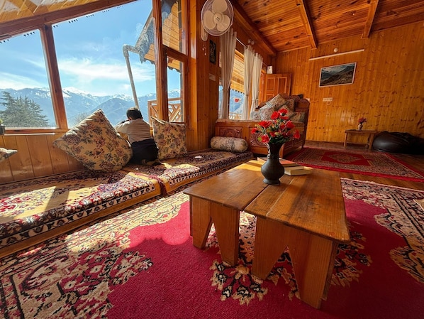 A cozy wooden cabin with a large window, a fan, a sofa with cushions, a table with a vase of flowers, and a rug.