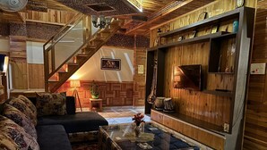 A cozy living room with a dark sofa, a glass coffee table, a wooden staircase, and a wall-mounted shelf with decorative items. - Cafe Saffron Sips & Stays (Kullu)