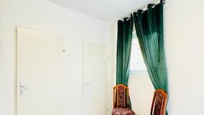 Een kamer met een glazen tafel, twee houten stoelen, een witte deur en groene gordijnen.