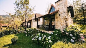 Ein Steinhaus mit Veranda, umgeben von üppiger Vegetation und blühenden Pflanzen.