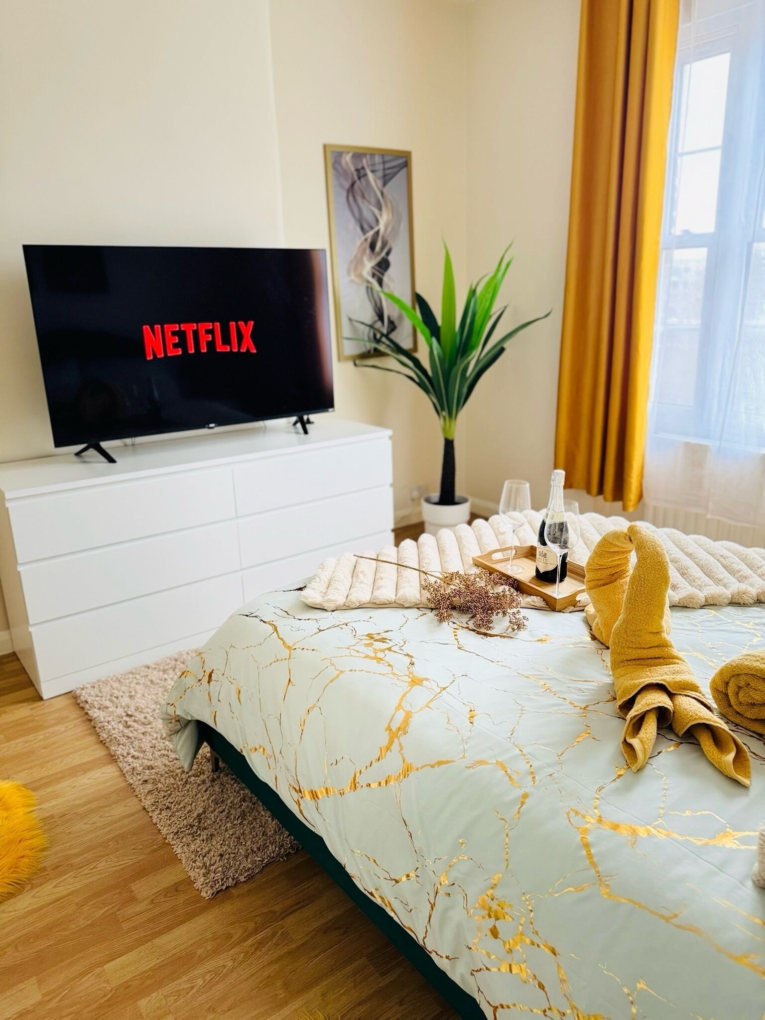 Ein Schlafzimmer mit einem Bett, einem Fernseher, der 'NETFLIX' anzeigt, einer Pflanze und einem Fenster mit Vorhängen.