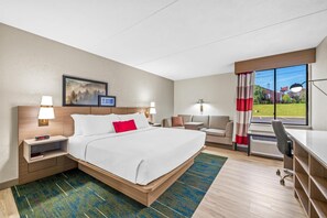 Ein Hotelzimmer mit Bett, Nachttischen, Schreibtisch, Stuhl, einer Couch und einem Fenster mit Blick auf Grünflächen.