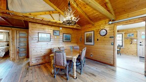 Ein Holzblockhaus-Innenraum mit Esstisch, Stühlen, einer Chandelüster, gerahmten Bildern und einer Wanduhr.