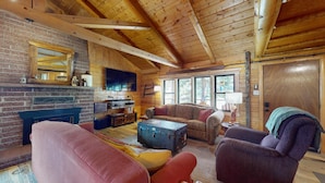 A cozy log cabin interior with a fireplace, wooden ceiling beams, a large TV, and a comfortable seating area.