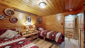 Ein blockhaushaltsähnliches Zimmer mit Holzwänden und Holzdielen, zwei Betten mit gemusterten Betttextilien, ein Schreibtisch mit Lampen und ein an der Wand hängender Traumfänger.