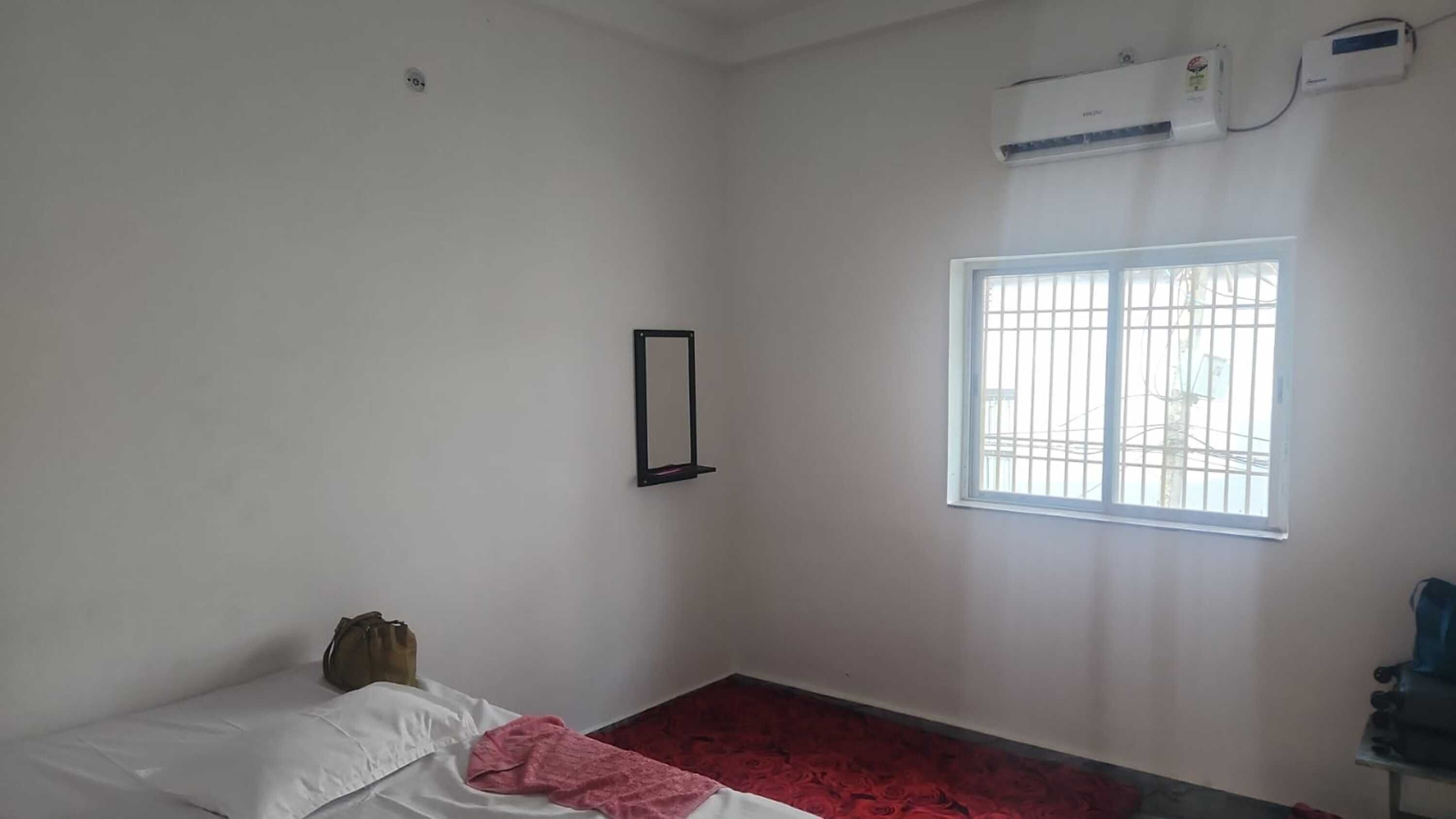 Una habitación con cama, ventana, aire acondicionado y un espejo fijado a la pared.