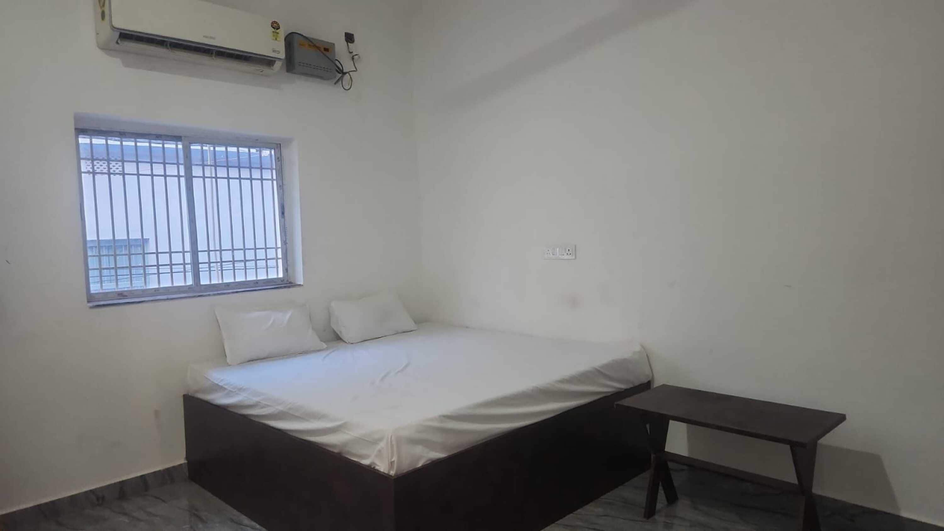 A bedroom with a bed, a window with bars, a wall-mounted air conditioner, and a small table.