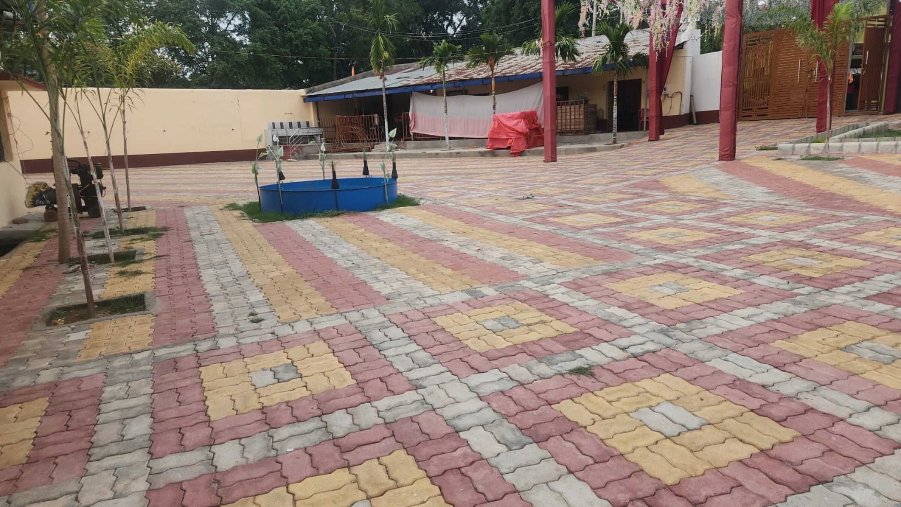 Un patio empedrado con un contenedor azul, árboles y un edificio con un pilar rojo.